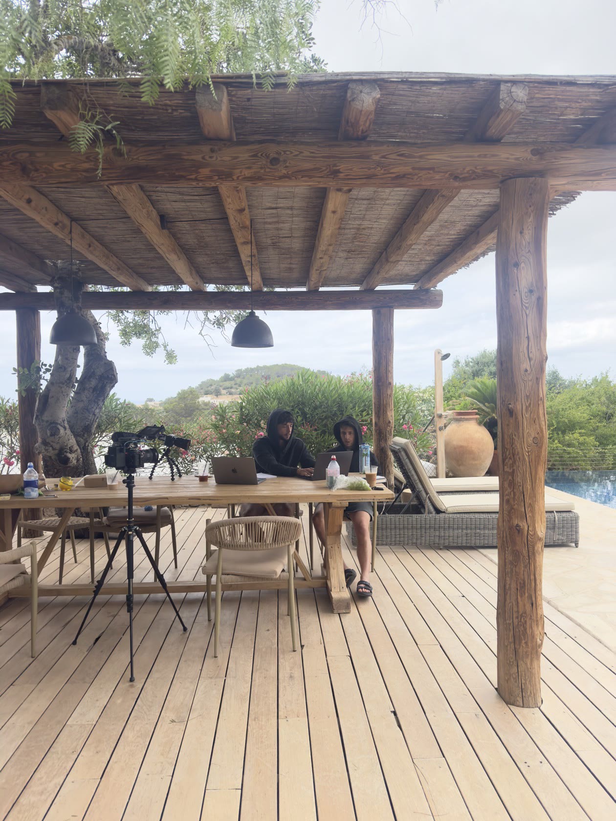 Two crew members building on laptops under a wooden pergola beside the villa's infinity pool, a cinema camera on a tripod in the foreground